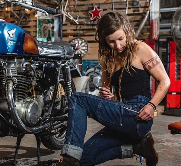 A woman with long dreadlocks sits in a garage, working on a vintage motorcycle. The setting is rustic with various tools and motorcycle parts around. She wears jeans, a black tank top, and brown boots, focused on her task. Image courtesy of Nathalie Dupré Photography.